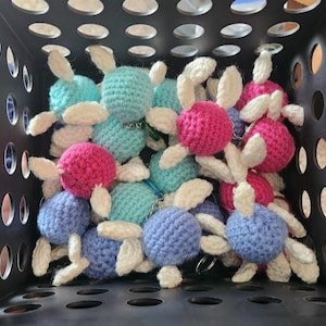 May include: A collection of handmade crochet bunny keychains in a black storage bin. The keychains are in various colors, including blue, pink, and teal, with white accents. Each bunny has a small metal ring for attaching keys.