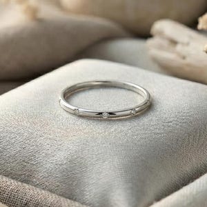 May include: A delicate silver ring with small, clear gemstones set into the band. The ring is displayed on a soft, gray cushion, surrounded by natural elements like stones and dried flowers, creating a serene and elegant composition.