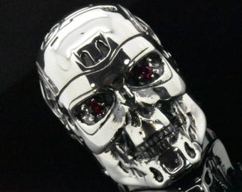 May include: A polished silver skull ring with red gemstone eyes. The ring is highly detailed, with a realistic skull design. The skull's features are sharply defined, and the ring has a shiny, reflective surface. The ring is a gothic or biker-style accessory.