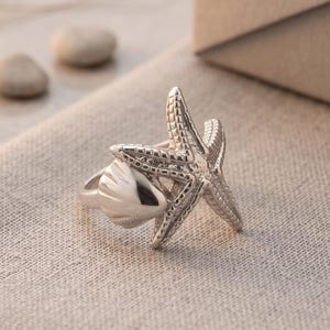May include: A silver-toned ring featuring a textured starfish design overlapping a smooth, polished seashell element. The metal band is connected to the starfish and shell motifs, creating a cohesive piece of jewelry with a metallic finish.