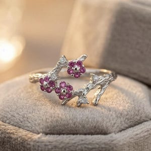 Könnte beinhalten: Ein silberfarbener Ring mit einem floralen Astdesign. Der Ring hat drei Gruppen kleiner, leuchtend rosa Blüten und einige kleine, klare Edelsteine. Der Ring liegt auf einer weichen, grauen Oberfläche.