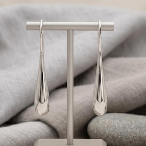 May include: A pair of silver teardrop earrings hanging from a silver display stand. The earrings have a smooth, polished finish and a long, slender shape. The stand is gray and sits on a white surface, with gray fabric and stones in the background.