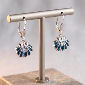 May include: Silver-tone seashell earrings with a dark blue enamel finish and a small white pearl. The earrings are displayed on a silver-coloured jewellery stand.