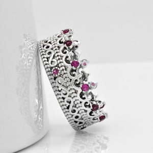 May include: A silver crown ring with an ornate filigree design and small, round, red gemstones. The ring is leaning against a white surface, showcasing its intricate details and regal aesthetic. The ring's design is reminiscent of a royal crown.