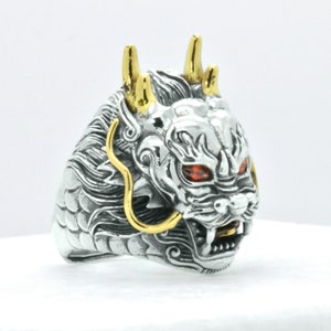 May include: A silver dragon ring with gold accents and red eyes. The dragon's head is facing forward with its mouth open and teeth showing. The ring is detailed with scales and intricate designs.