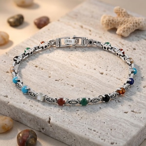 May include: A sterling silver bracelet featuring a series of small, round, multi-colored gemstones and dark, textured metal accents. The clasp is clearly marked with the number 925, indicating sterling silver composition. The design is circular.