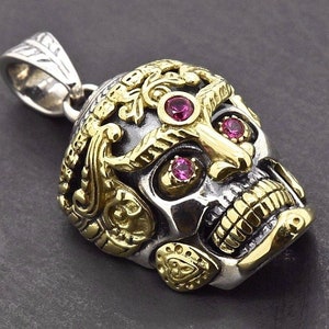 Op de afbeelding: Zilveren schedel hanger met gouden accenten en drie roze edelstenen. De schedel heeft ingewikkelde details, waaronder sierlijke gouden filigraan en een gedetailleerde kaaklijn. De hanger heeft een zilveren balein.