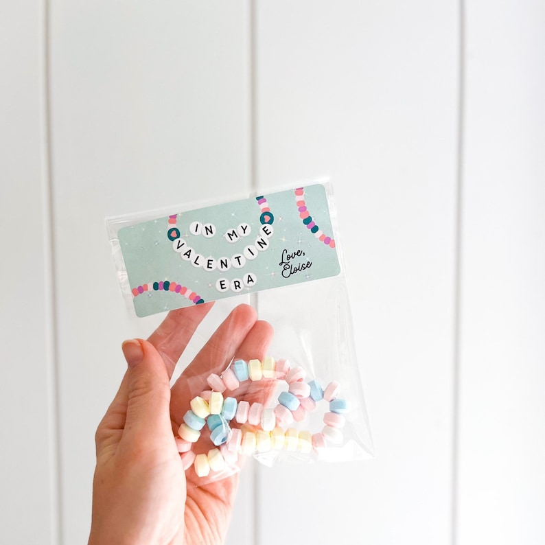 May include: A clear plastic bag containing pastel-colored candy necklaces. The bag has a card with the text "IN MY VALENTINE ERA" and "Love, Eloise" on a light green background. The candy necklaces are in shades of pink, blue, yellow, and white.