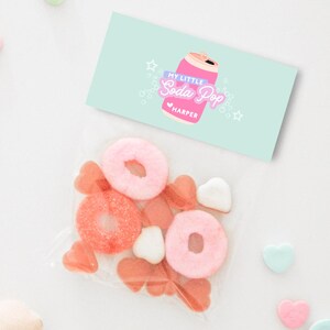 May include: A clear cellophane bag filled with pink and red candies, including ring-shaped and heart-shaped sweets. A light blue card with the text "My Little Soda Pop" and a soda can illustration is placed above the bag.