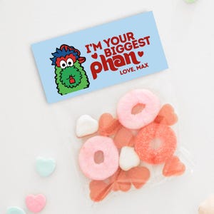 May include: A light blue card with the text "I'M YOUR BIGGEST phan" and a cartoon character. A clear bag contains pink and orange candies. Scattered around are heart-shaped candies in pink and blue.