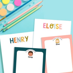 Op de afbeelding: Briefpapier set met gepersonaliseerde notitieblokken met de namen "HENRY" en "ELOISE" in kleurrijke letters. De set bevat potloden, waterverf en extra notitieblokken met geïllustreerde karakters. De achtergrond is lichtblauw.