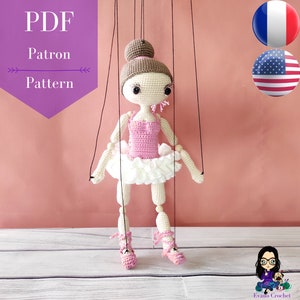 May include: A crochet ballerina puppet with pink and white clothing, brown hair, and black eyes. The puppet is hanging from strings and is ready to dance. The image includes the text "PDF Patron Pattern".
