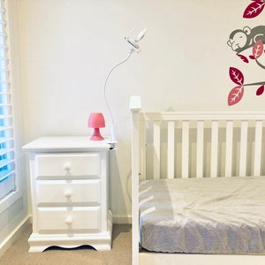 May include: A white cot with a white mattress and a white bedside table with three drawers. A pink lamp sits on the bedside table. A white lamp with a flexible arm is mounted on the wall. A grey monkey is sleeping on a tree branch with red leaves.