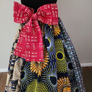 May include: A vibrant skirt with a large red bow. The skirt features a patchwork design with bold patterns in blue, yellow, and brown. The bow has a white geometric pattern on a red background. The skirt is displayed on a black mannequin.