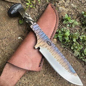 May include: A handcrafted knife with a textured, multi-colored blade and a dark, ornate handle. The knife rests on a brown leather sheath, set against a natural outdoor backdrop. The blade has a unique pattern and the handle has a detailed design.