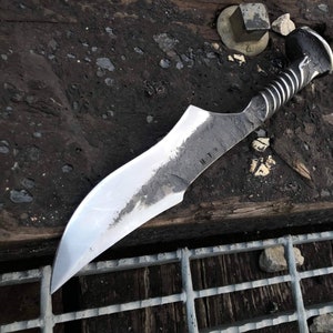 May include: A handmade knife with a silver blade and a black handle. The blade is curved and has a pointed tip. The handle is made of a twisted metal and has a nail head at the end. The knife is resting on a wooden surface.