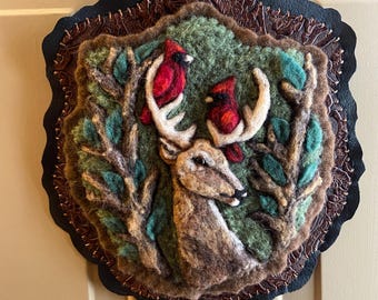 May include: A felted wall hanging featuring a deer with antlers, two red cardinals, and green foliage. The artwork is framed with a dark leather border and a brown, textured inner border. The overall design has a rustic, nature-inspired theme.