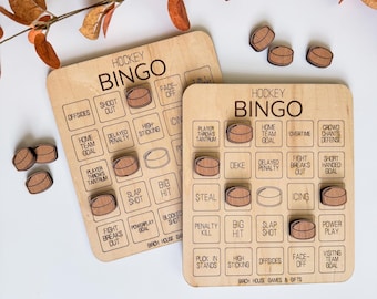 May include: Two wooden hockey bingo boards with wooden hockey puck game pieces. The boards have printed squares with hockey terms like "Offsides" and "Penalty Kill". The text "Hockey Bingo" is printed at the top of each board.