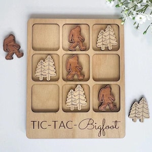 May include: A wooden tic-tac-toe game with the words "Tic-Tac-Bigfoot" engraved on the board. The game pieces are wooden and feature images of Bigfoot and pine trees.