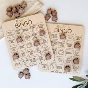May include: Two wooden Reality TV Bingo game boards with matching wooden TV-shaped game pieces. The boards feature phrases like "Crying with Mascara" and "Dramatic Confession". The game is from Birch House Games & Gifts.