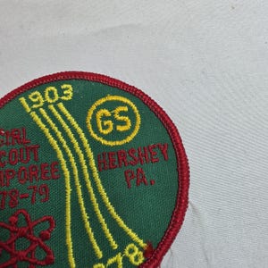 May include: A circular Girl Scout patch with a green background and red border. The patch features yellow embroidered text and design elements, including the year "1903" and the words "Hershey PA." A yellow circle with "GS" is also present.