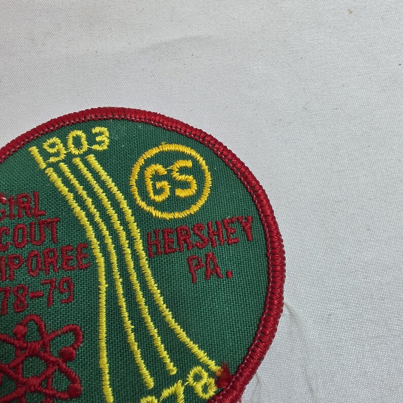 May include: A circular Girl Scout patch with a green background and red border. The patch features yellow embroidered text and design elements, including the year "1903" and the words "Hershey PA." A yellow circle with "GS" is also present.