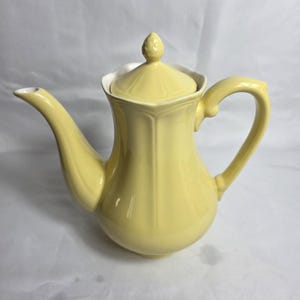 May include: A pale yellow ceramic teapot with a matching lid. The teapot has a curved spout and a handle. The lid is topped with a small, decorative finial. The teapot has a white rim and a bulbous body.