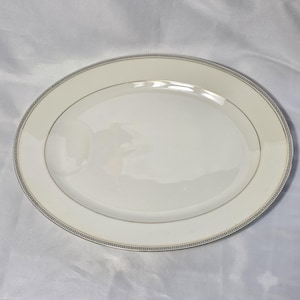 May include: An oval-shaped, off-white serving platter with a gold trim and a decorative border. The border features a repeating geometric pattern in gray and black. The platter has a smooth, glossy surface.