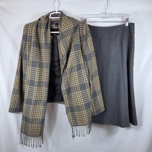 May include: A gray and tan houndstooth pattern jacket with a matching gray skirt. The jacket has a fringe scarf and is hanging on a white hanger. The skirt is a solid gray color and is hanging on a clear hanger.