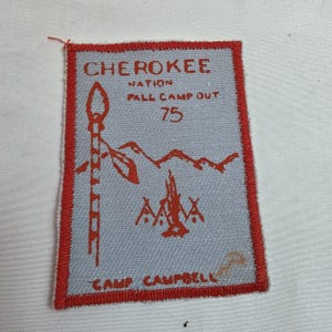 May include: A rectangular patch with a red border and a light blue center. The patch features the text "CHEROKEE NATION FALL CAMP OUT 75" in red. It also includes red illustrations of a spear, mountains, a campfire, and the text "CAMP CAMPBELL".