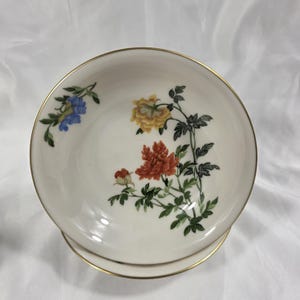 May include: A white porcelain bowl with a gold rim, featuring a floral design in shades of blue, yellow, and orange. The flowers are accompanied by green leaves and stems. The bowl is resting on a matching saucer.