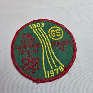 May include: A round, embroidered Girl Scout Camporee patch. The patch is green with red trim and features the text "Girl Scout Camporee 1978-79 Hershey PA." and the years 1903 and 1978. A yellow atomic symbol and GS logo are also present.