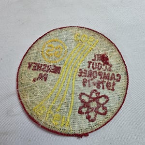 May include: A round, embroidered patch with a red border. The patch features yellow and red stitching with text and graphics. The text includes "23" and "Hershey, PA". The patch also has a flower and a series of yellow lines.