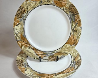 May include: Two white ceramic plates with a decorative border featuring a pattern of leaves and pinecones in shades of brown, tan, and gray. The plates are stacked, showcasing the design.