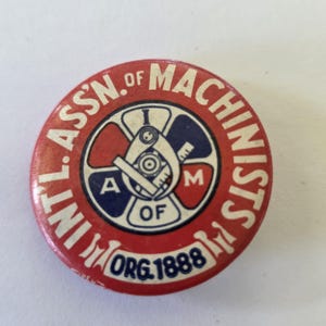 May include: A round, red button with white text reading "INT'L ASSN. OF MACHINISTS" around the edge. The center features a blue and white emblem with the letters "A" and "M" and the text "ORG. 1888".