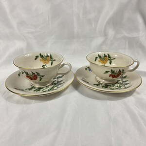 May include: Two cream-colored teacups and saucers. Each cup and saucer features a floral design with red, yellow, and green accents. The rims are trimmed with gold. The teacups have small handles.