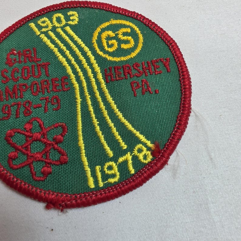 May include: A green Girl Scout Camporee patch with red and yellow embroidered details. The patch features the text "Girl Scout Camporee 1978-79" and "Hershey PA." along with the years 1903 and 1978. A red atom symbol is also present.