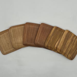 May include: Five wooden coasters in shades of brown, arranged in a fan shape. The coasters have rounded corners and a slightly raised edge. The wood grain is visible, adding texture to the coasters.