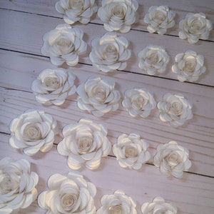 May include: A collection of 17 ivory paper roses with a subtle shimmer. The roses are arranged in rows on a light wood surface.