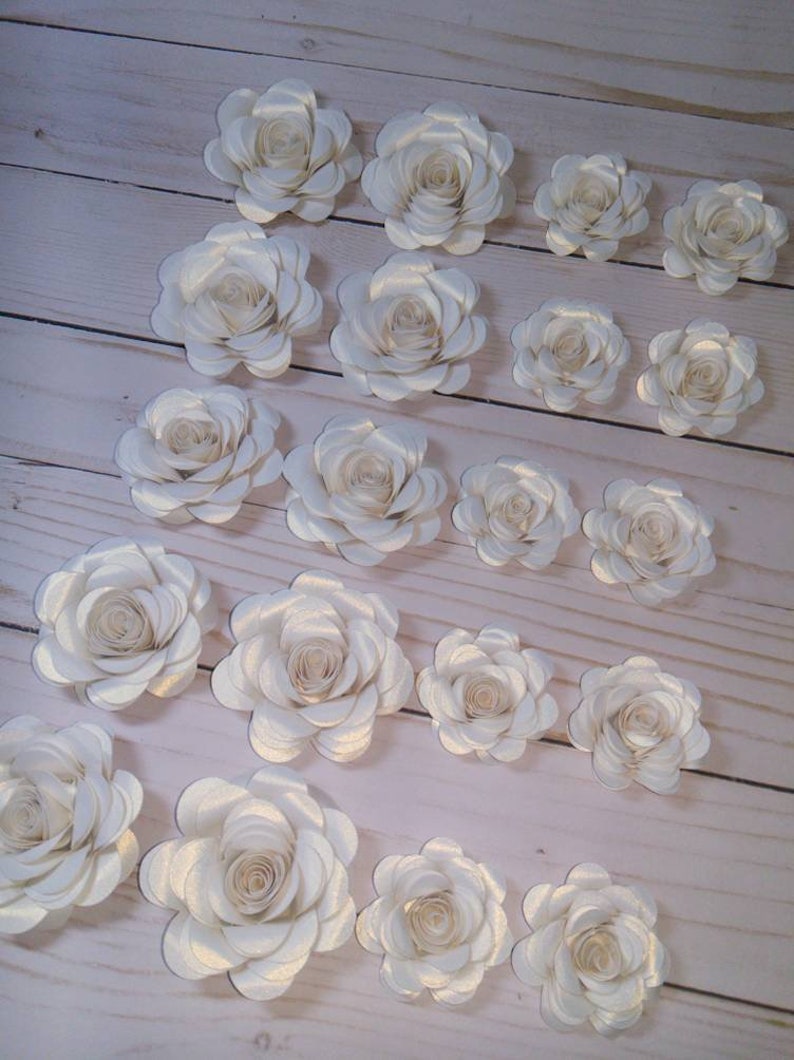 May include: A collection of 17 ivory paper roses with a subtle shimmer. The roses are arranged in rows on a light wood surface.