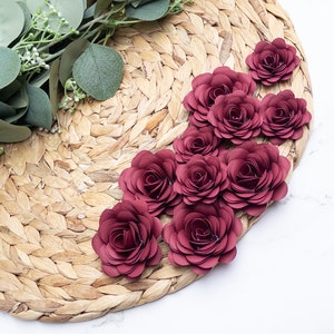 May include: A close-up of a woven straw placemat with eight burgundy paper roses arranged in a semi-circle. The roses are layered and have a delicate, ruffled appearance.