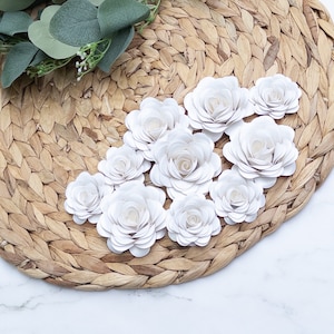 May include: A collection of white paper flowers arranged on a woven straw placemat. The flowers are made of multiple layers of paper, creating a delicate and layered effect.
