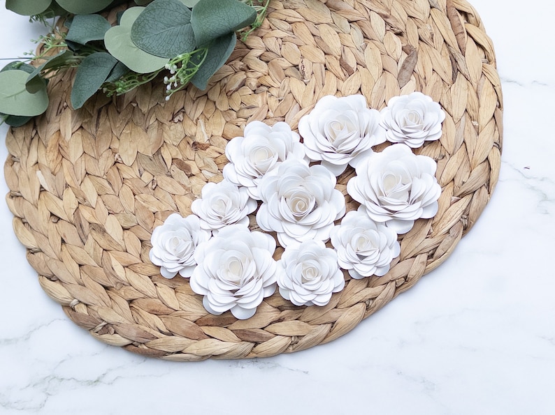 May include: A collection of white paper flowers arranged on a woven straw placemat. The flowers are made of multiple layers of paper, creating a delicate and layered effect.