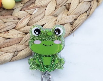 May include: A green glitter frog with a smiling face and pink cheeks is attached to a clear plastic retractable badge holder with a silver clip.