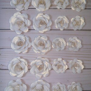 May include: A set of 20 white paper flowers with a subtle shimmer. The flowers are arranged in rows, with the largest flowers at the top and the smallest flowers at the bottom. The flowers are all made with the same technique, with multiple layers of paper that are curled and layered to create a realistic look.