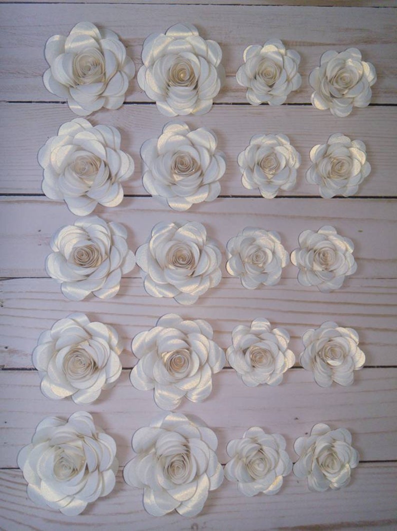 May include: A set of 20 white paper flowers with a subtle shimmer. The flowers are arranged in rows, with the largest flowers at the top and the smallest flowers at the bottom. The flowers are all made with the same technique, with multiple layers of paper that are curled and layered to create a realistic look.