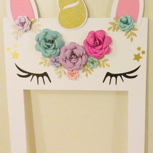 May include: White unicorn photo booth frame with pink ears, a gold glitter horn, and paper flowers in shades of pink, purple, and blue. The frame has a cutout for a photo and the text "Brisecida's 6th Birthday" at the bottom.
