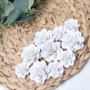 May include: A collection of white paper roses arranged on a woven straw placemat. The roses are made of multiple layers of paper, giving them a delicate and realistic appearance.