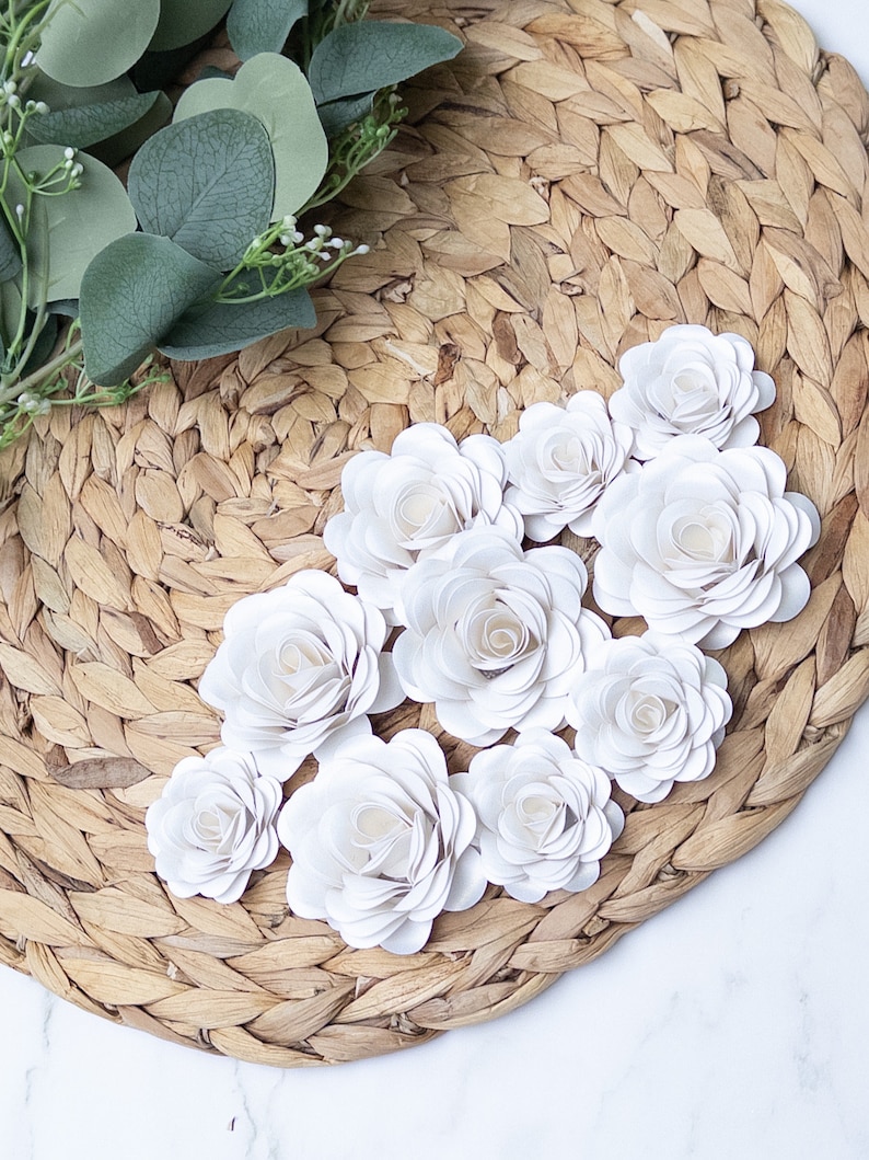 May include: A collection of white paper roses arranged on a woven straw placemat. The roses are made of multiple layers of paper, giving them a delicate and realistic appearance.
