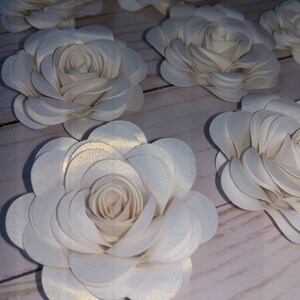 May include: A collection of white paper flowers with a subtle shimmer. The flowers are layered and have a delicate, feminine appearance.